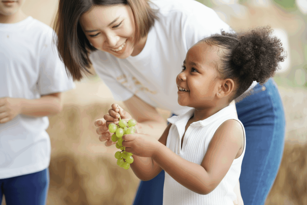 A young child holding a bunch of green grapes smiles as an adult woman leans in, both appearing happy. Another person in a white shirt is partially visible on the left.