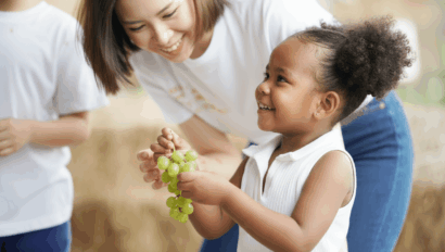 A young child holding a bunch of green grapes smiles as an adult woman leans in, both appearing happy. Another person in a white shirt is partially visible on the left.