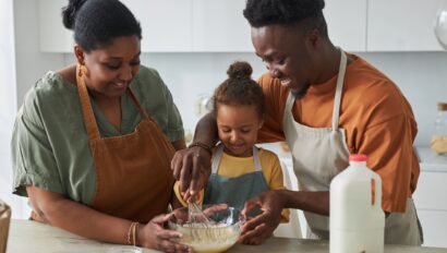 Three people, two adults and a child, are standing in a kitchen mixing ingredients in a bowl, wearing aprons and smiling. A bottle of milk and eggs are on the counter.